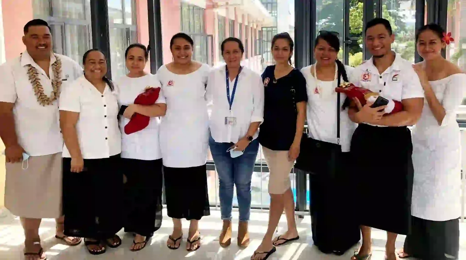 Nine people, appearing to be staff or employees, stand together. They are all smiling and most are wearing white shirts with black accents, and black skirts or pants. Some have leis around their necks.