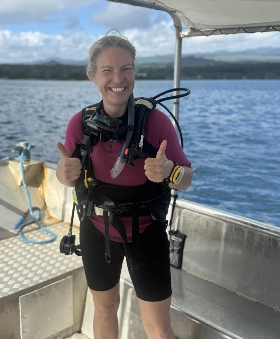 A smiling female scuba diver stands on the edge of a boat, giving a thumbs-up. The diver is wearing a full scuba setup, including a mask, regulator, BCD, and tank. Behind them is a calm, blue ocean with lush green hills visible in the distance.