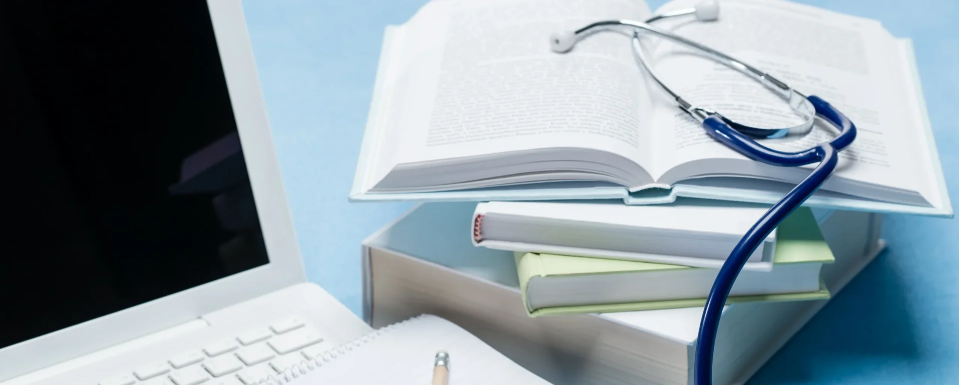 A stethoscope draped over an open book and a laptop, resting on a stack of books.