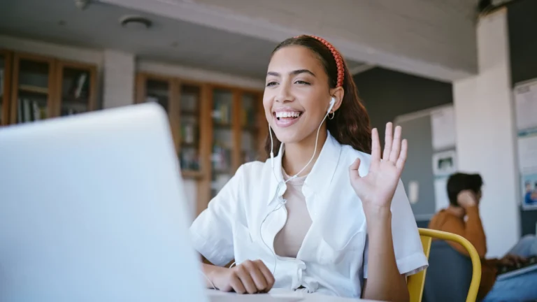 Smiling young woman wearing earphones, sitting at a desk and waving during a video call on her laptop in a modern study space