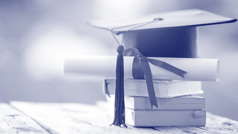 Graduation cap with tassel and a rolled diploma tied with a ribbon placed on top of a stack of books
