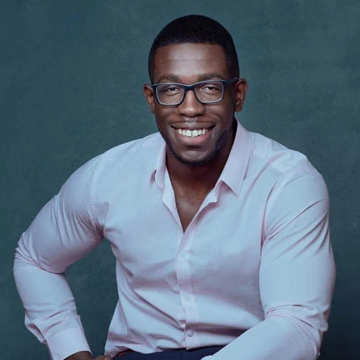A smiling man with dark hair and glasses looks directly at the viewer. He is wearing a light pink button-down shirt and appears to be sitting or leaning back. The background behind him is a muted teal color.