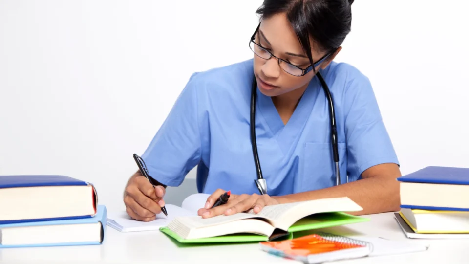 Medical student in blue scrubs with a stethoscope studies at a desk, writing notes in an open textbook surrounded by other books and notebooks