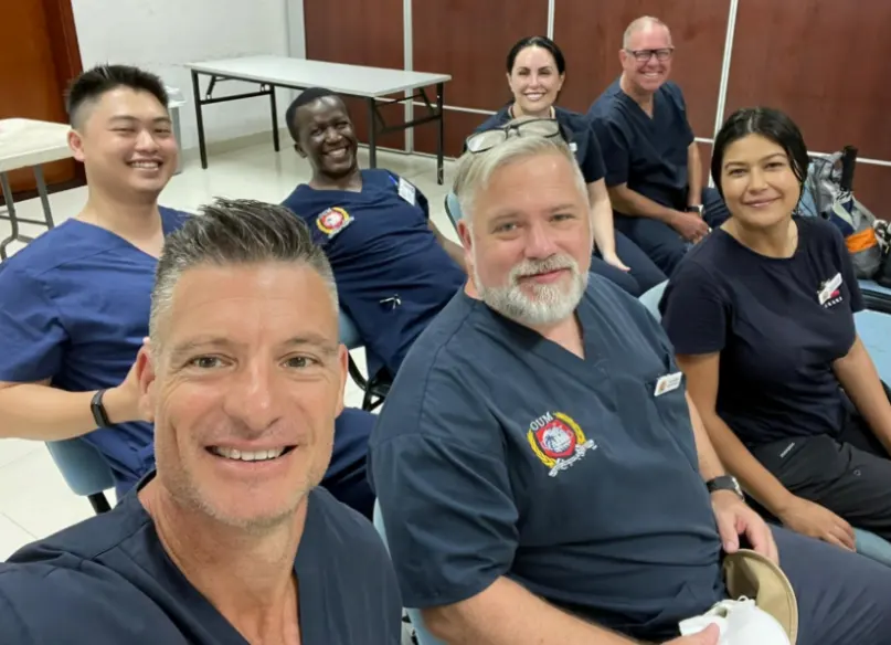 A group of eight doctors, including both men and women, are smiling and posing for a group photo. They are wearing dark blue scrubs and are standing in a room with a table in the background.