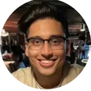 A smiling young man with dark wavy hair and eye glasses wearing a tan colored shirt