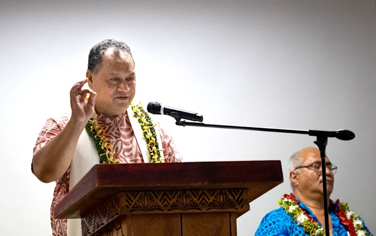 A man in a floral shirt is speaking at a podium. Another man is standing to his right, also wearing flowers.